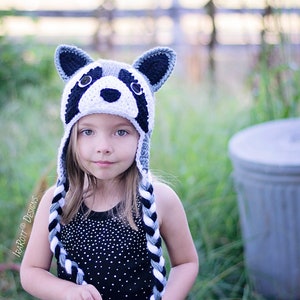 May include: A young girl wearing a gray and black crocheted raccoon hat with black and white braided ties. The hat has a white crocheted face with black eyes and a black nose.