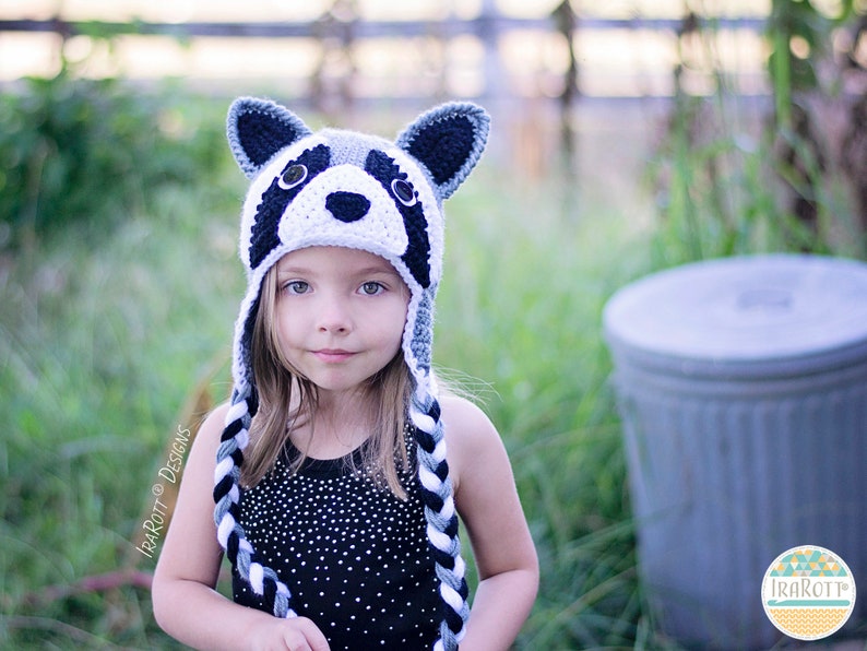 May include: A young girl wearing a gray and black crocheted raccoon hat with black and white braided ties. The hat has a white crocheted face with black eyes and a black nose.