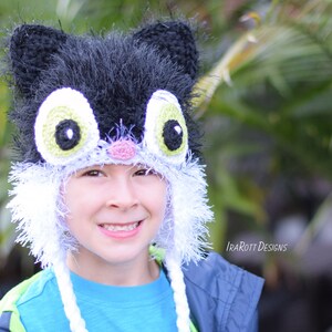 May include: A handmade black and white cat hat with large, expressive eyes and ear details. The hat features a fluffy texture and is designed to be worn on the head. The hat has a playful and whimsical design, suitable for costume or everyday wear.