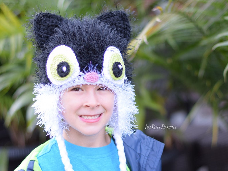 May include: A handmade black and white cat hat with large, expressive eyes and ear details. The hat features a fluffy texture and is designed to be worn on the head. The hat has a playful and whimsical design, suitable for costume or everyday wear.