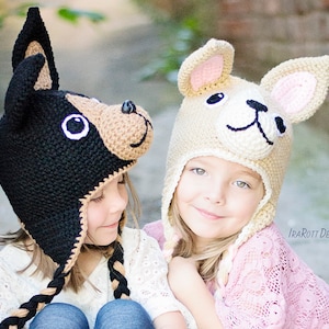 May include: Two children wearing crocheted dog hats. The girl on the left is wearing a black and brown hat with floppy ears. The girl on the right is wearing a beige hat with pink ears. Both hats have embroidered faces.