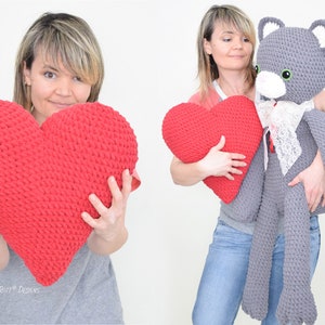 May include: Two women are holding crocheted items. One woman is holding a large grey crocheted cat with white accents. The other woman is holding a red crocheted heart-shaped cushion.