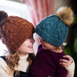 May include: Two people wearing knitted hats with pom poms. The person on the left is wearing a brown and orange hat, and the person on the right is wearing a teal blue hat.