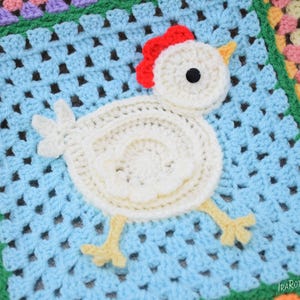Pode incluir: Um cobertor de croch&ecirc; de quadrados da av&oacute; com uma aplica&ccedil;&atilde;o de frango de croch&ecirc; branco no centro. O frango tem uma crista vermelha, olhos pretos e p&eacute;s amarelos.
