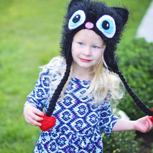 May include: A black cat hat with large blue eyes, a pink nose, and long black braids ending in red hearts. The hat is worn by a young person with blonde hair, wearing a blue and white patterned dress. The hat is handmade.