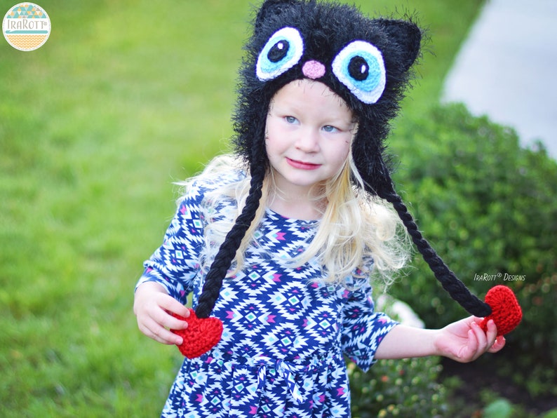 May include: A black cat hat with large blue eyes, a pink nose, and long black braids ending in red hearts. The hat is worn by a young person with blonde hair, wearing a blue and white patterned dress. The hat is handmade.