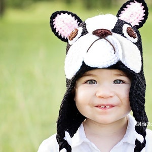 May include: A child wearing a black and white crocheted hat shaped like a Boston Terrier dog. The hat has pink ears and brown eyes. The child is smiling.