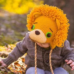 May include: A child wearing a crocheted lion hat with a brown sweater and blue jeans. The hat has green eyes and a brown nose.