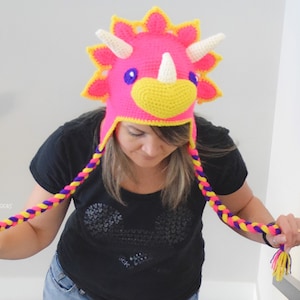 Puede incluir: Un sombrero de triceratops de ganchillo hecho a mano en rosa brillante, amarillo y blanco. El sombrero presenta una cabeza de dinosaurio con cuernos y un pico amarillo. Cuelgan borlas trenzadas en morado, amarillo y rosa. El sombrero es un accesorio divertido.