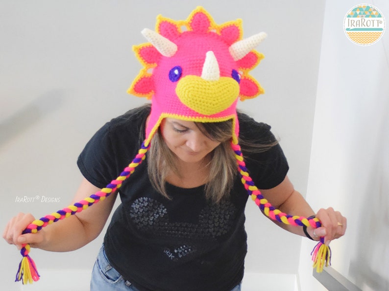 Puede incluir: Un sombrero de triceratops de ganchillo hecho a mano en rosa brillante, amarillo y blanco. El sombrero presenta una cabeza de dinosaurio con cuernos y un pico amarillo. Cuelgan borlas trenzadas en morado, amarillo y rosa. El sombrero es un accesorio divertido.