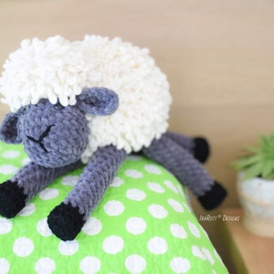 May include: A crocheted plush sheep toy with a white fluffy body and gray velvet legs and head. The sheep is lying on a green and white polka dot pillow.