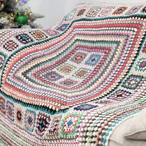 Può includere: Una coperta all'uncinetto colorata con un grande motivo ovale al centro. La coperta è realizzata con una varietà di filati di diversi colori, tra cui rosa, blu, verde e marrone. La coperta è drappeggiata su un divano bianco.