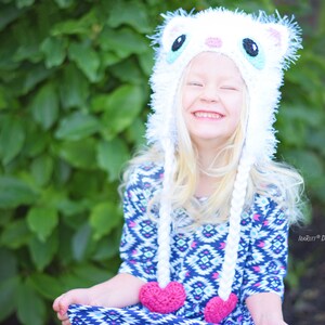 May include: A white crocheted cat hat with long braids and pink heart accents. The hat features a cat face design with black and blue eyes. The braids hang down with pink hearts at the ends. The hat is worn by a person with blonde hair.