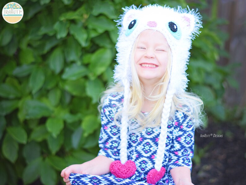 May include: A white crocheted cat hat with long braids and pink heart accents. The hat features a cat face design with black and blue eyes. The braids hang down with pink hearts at the ends. The hat is worn by a person with blonde hair.