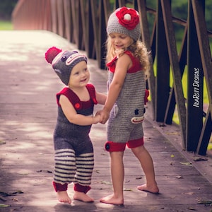 Puede incluir: Dos niños con trajes de mono de punto gris y rojo a juego con gorros. La niña lleva una flor roja en el gorro. Se están dando la mano y están de pie en un puente de madera.