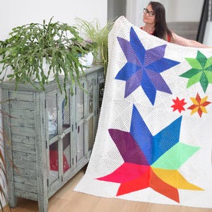 May include: A white crocheted blanket featuring colorful star designs in shades of blue, purple, green, red, orange, and yellow. The blanket is displayed in a well-lit room with a wooden cabinet and plants.