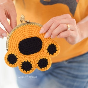 May include: A crocheted coin purse in the shape of a paw, held by a person. The purse is primarily orange with a black paw pad and black circles representing the paw's toes. The purse has a metal clasp and the text "IraRott Designs".