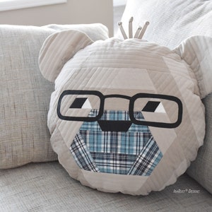 以下が含まれることがあります： ベージュのキルティング枕。クマの顔、黒い眼鏡、青と白の格子柄の鼻。枕はベージュのソファの上にある。