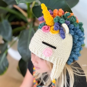 May include: A white crocheted unicorn hat with a rainbow mane and horn. The hat has a black eye and a pink cheek. The hat is being worn by a child.