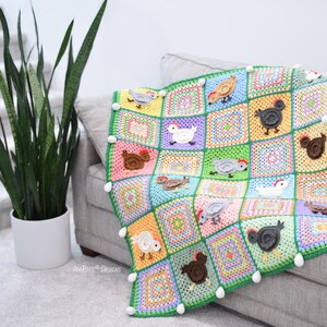 Pode incluir: Uma manta de croch&ecirc; colorida com um design de patchwork que apresenta quadrados de v&aacute;rias cores e galinhas aplicadas. A manta &eacute; decorada com pompons brancos ao redor das bordas.