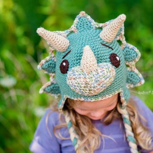 Puede incluir: Un gorro de ganchillo hecho a mano con forma de triceratops. El gorro es principalmente de color verde azulado con cuernos y detalles beige, ojos marrones y un hocico beige. El gorro tiene orejeras y lazos trenzados. El fondo es verde borroso.