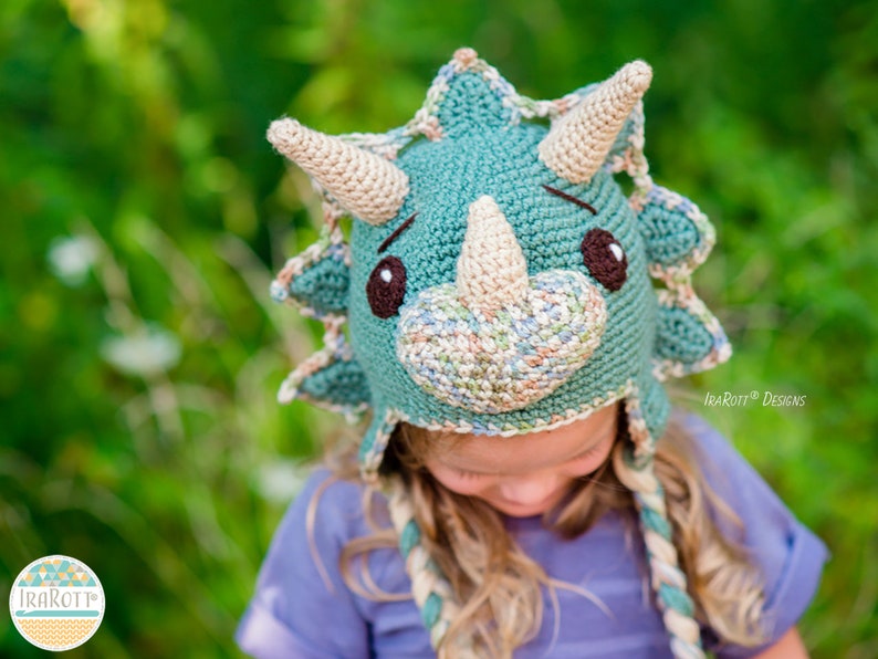 Puede incluir: Un gorro de ganchillo hecho a mano con forma de triceratops. El gorro es principalmente de color verde azulado con cuernos y detalles beige, ojos marrones y un hocico beige. El gorro tiene orejeras y lazos trenzados. El fondo es verde borroso.