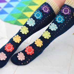 May include: A pair of navy blue crochet knee-high socks adorned with colorful crochet flowers. The flowers are in shades of pink, red, yellow, green, blue, and purple. The top of the socks is a beige color with a yellow and green tie.
