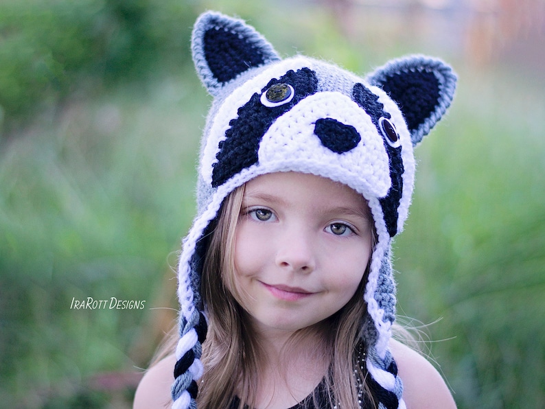 May include: A young person wearing a gray and white crocheted raccoon hat with black eyes and a black nose. The hat has long, braided ties.