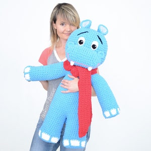 May include: A large blue crocheted hippopotamus wearing a red scarf. The hippopotamus is being held by a person.