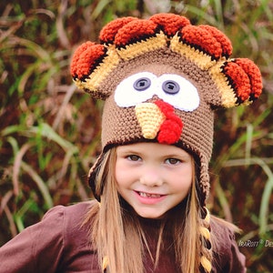 May include: A child wearing a brown, red, and yellow crocheted turkey hat with white felt eyes and a red felt wattle. The hat has a brown crocheted brim and ear flaps.