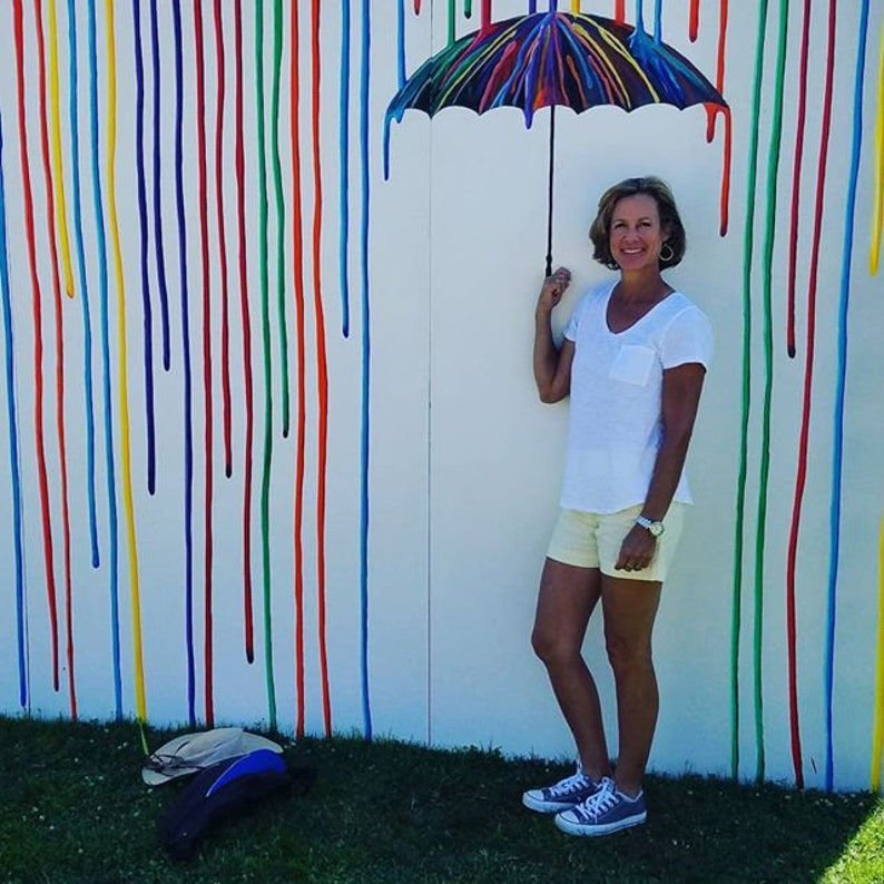May include: A woman stands in front of a white wall with colorful paint streaks. She is holding a black and white umbrella over her head. She is wearing a white t-shirt, light yellow shorts, and gray sneakers.