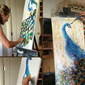 May include: A woman paints a peacock on a canvas. The peacock is blue with a green and gold tail. The woman is wearing a green apron and a grey shirt.