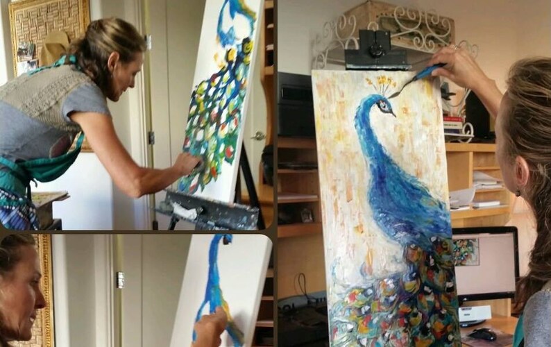 May include: A woman paints a peacock on a canvas. The peacock is blue with a green and gold tail. The woman is wearing a green apron and a grey shirt.