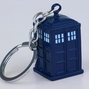 Könnte beinhalten: Ein dunkelblauer TARDIS-Schlüsselanhänger mit einer silberfarbenen Metallkette und einem Schlüsselring. Die Miniatur-Polizeibox hat weiße Fensterdetails und eine Schlaufe oben für die Kette.