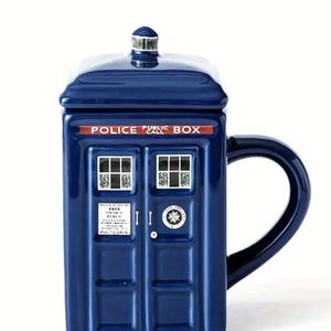 Könnte beinhalten: Eine marineblaue Keramiktasse in Form einer britischen Polizeirufzelle, mit passendem Deckel. Auf der Tasse steht "POLICE PUBLIC CALL BOX" und sie hat zwei Fenster und ein Türendesign. Der Griff ist in Marineblau gehalten.