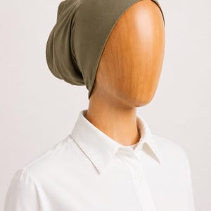 May include: An olive green beanie-style head covering is displayed on a wooden mannequin head. The head covering is made of a soft fabric. The mannequin is wearing a white collared shirt.