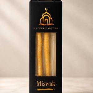 May include: A black box with the text "SUNNAH GOODS" and "Miswak" in gold lettering. The box contains three light-colored sticks visible through a clear window. The top of the box features a gold emblem of a mosque and crescent moon.