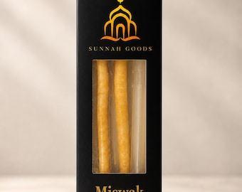 May include: A black box with the text "SUNNAH GOODS" and "Miswak" in gold lettering. The box contains three light-colored sticks visible through a clear window. The top of the box features a gold emblem of a mosque and crescent moon.