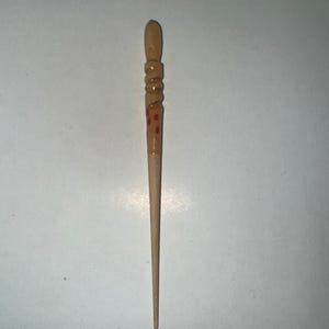 Könnte beinhalten: Ein Haarstab aus Holz mit hellbraunem Finish. Der obere Teil weist geschnitzte Details und kleine rote Punkte auf. Der Stab verjüngt sich zu einer Spitze. Ein schlichtes, aber elegantes Haaraccessoire.