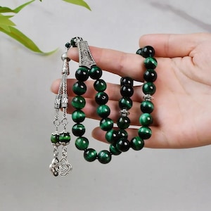 May include: Green tiger's eye prayer beads with silver accents and a tassel. The beads are strung on a silver cord and have a silver charm at the top.