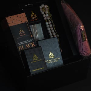 May include: A black gift box containing various items. Included are boxes of black seed oil and pure thimid oil, a beaded rosary, and a wrapped pink scarf. The boxes have the text "SUNNAH GOODS".