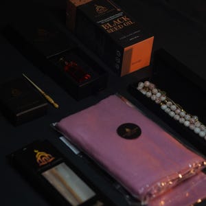 May include: An assortment of products, including a box of Black Seed Oil, a small bottle of red liquid, a pink scarf, and a beaded necklace, are arranged on a dark surface. The items are packaged in black boxes with gold accents and labels.