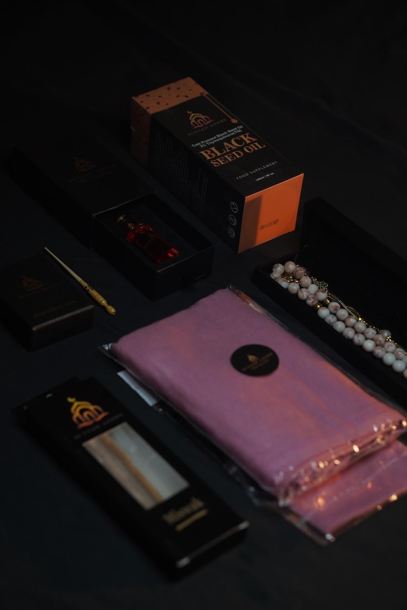 May include: An assortment of products, including a box of Black Seed Oil, a small bottle of red liquid, a pink scarf, and a beaded necklace, are arranged on a dark surface. The items are packaged in black boxes with gold accents and labels.