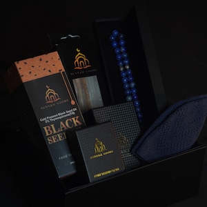 May include: A gift box containing various items, including a box of black seed oil, a blue beaded prayer beads, and a navy blue prayer cap. The boxes have the text "SUNNAH GOODS" and "BLACK SEED".