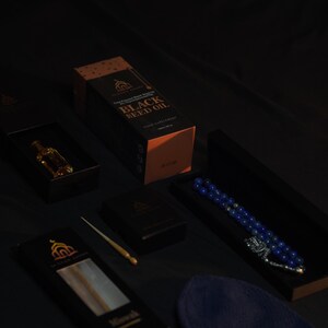 May include: An assortment of products, including a box of black seed oil, a small bottle, a prayer bead set, and a prayer cap. The items are arranged on a dark surface, with the black seed oil box prominently displayed. The box has gold lettering.