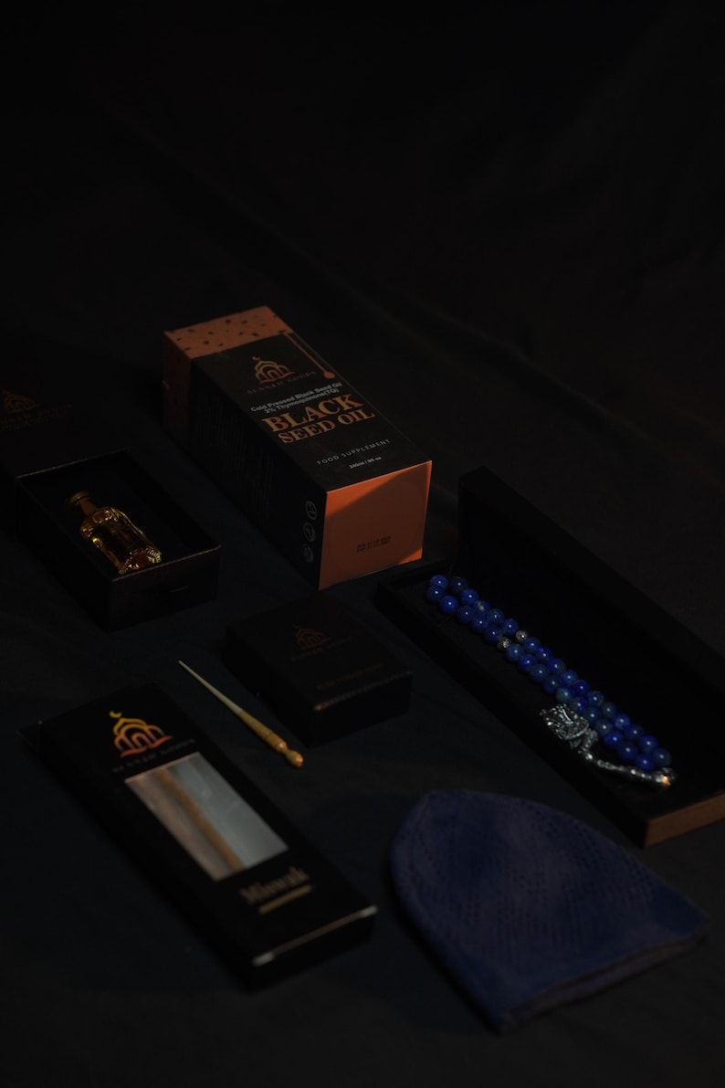 May include: An assortment of products, including a box of black seed oil, a small bottle, a prayer bead set, and a prayer cap. The items are arranged on a dark surface, with the black seed oil box prominently displayed. The box has gold lettering.