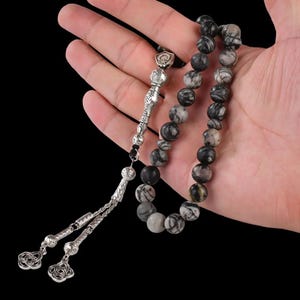 May include: A black and white prayer beads mala with a silver tassel and a silver charm. The beads are made of a stone with a marbled pattern.