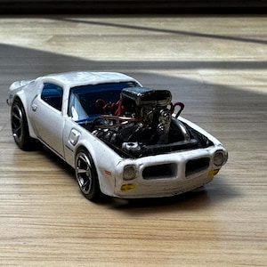 May include: A white toy car with a detailed engine, hood open. The car has black wheels and a blue interior. The engine is silver with red accents. The car is on a wooden surface.
