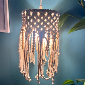Puede incluir: Una lámpara colgante de macramé hecha a mano. La pantalla de color blanco roto está intrincadamente anudada con una forma cilíndrica, adornada con cuentas de madera y largas borlas. La lámpara cuelga de un cable blanco contra una pared verde azulado.
