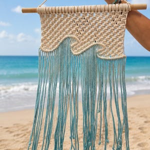 May include: A macrame wall hanging featuring a cream-colored woven top section with a wave-shaped design. The lower portion consists of long, light blue and cream-colored cotton cord fringe, all suspended from a horizontal wooden dowel rod.